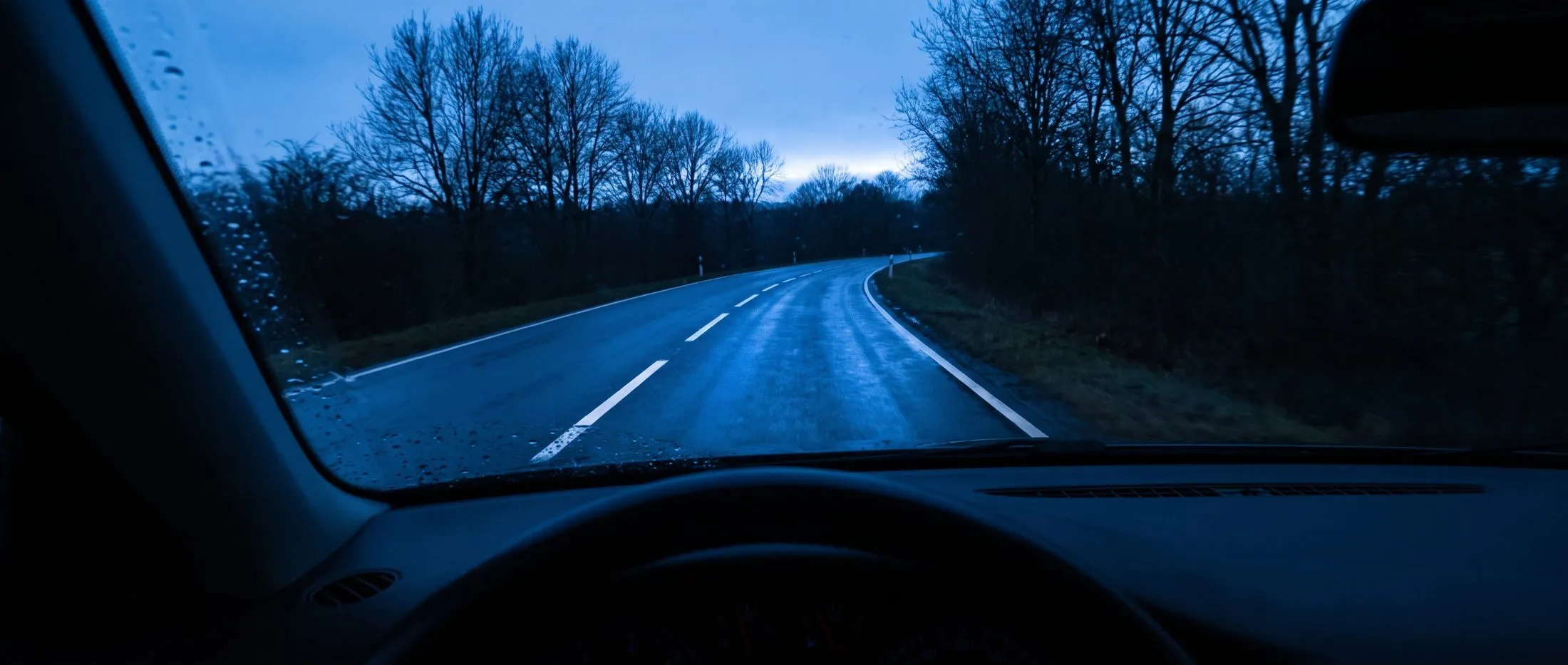 Blick durch die Windschutzscheibe auf eine nasse Landstraße in der blauen Stunde